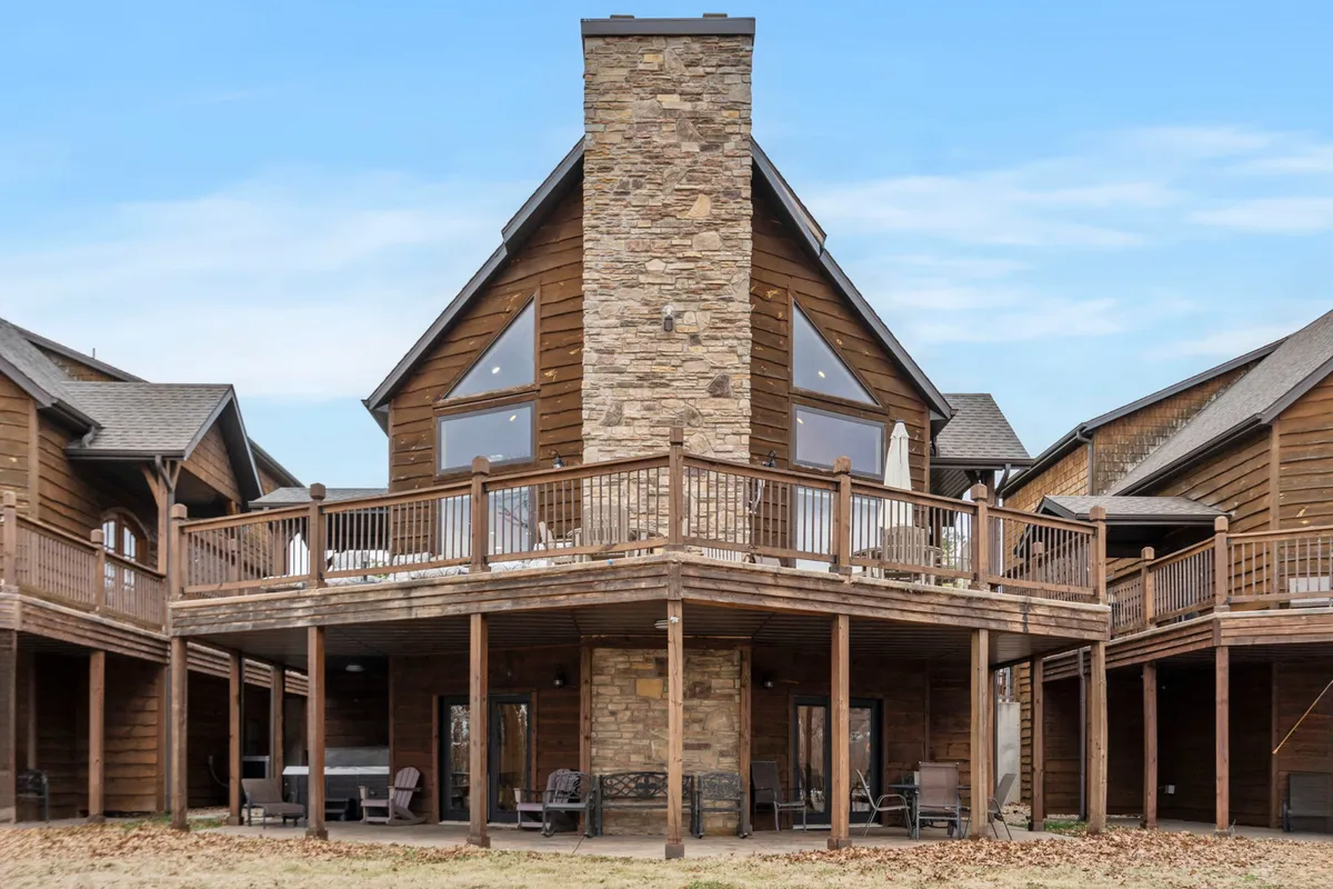 An exterior perspective of the property showcases an expansive wooden wrap-around deck featuring outdoor seating and a large stone chimney. The deck overlooks the lake with large windows hinting at the cozy interiors inside, inviting guests to enjoy both indoor comfort and outdoor serenity.