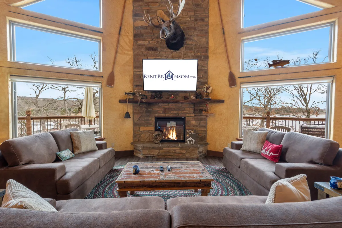 A spacious living room features a grand stone fireplace as the centerpiece, surrounded by comfortable sofas and a rustic coffee table. Floor-to-ceiling windows frame sweeping views of Table Rock Lake, inviting natural light and a sense of connection to the water beyond. A mounted deer head and wooden accents enhance the lodge-style, welcoming atmosphere.