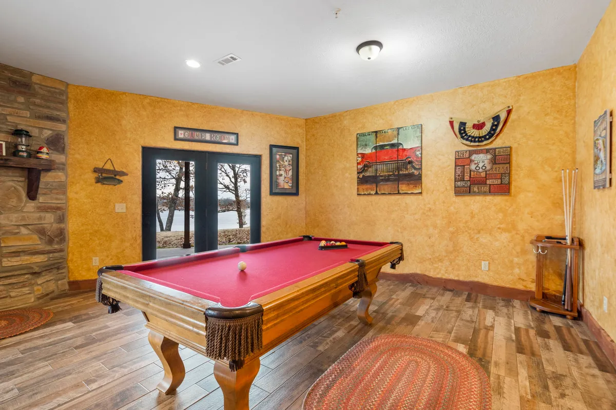 A stylish game and lounge area featuring a pool table at center, large windows with views toward the lake, a decorative wall art, and a cozy fireplace. The room invites both lively competition and relaxed conversation, bathed in natural light from the windows.