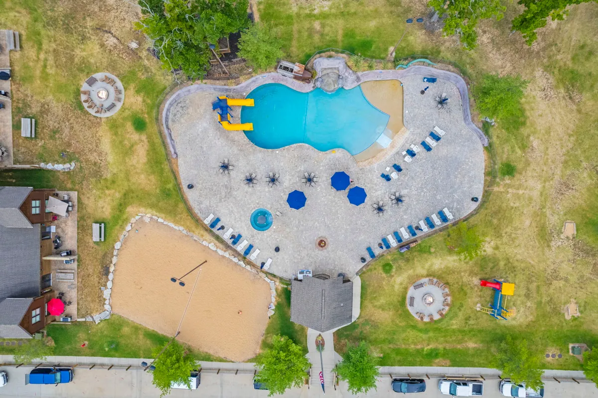 This image captures a vibrant exterior scene featuring a spacious swimming pool with a water slide, a sand volleyball court, several lounge chairs, and a hot tub. The area is bustling with activity, showing guests swimming, sunbathing, playing volleyball, and relaxing in the hot tub, all framed by a backdrop of lush greenery and sunny skies.