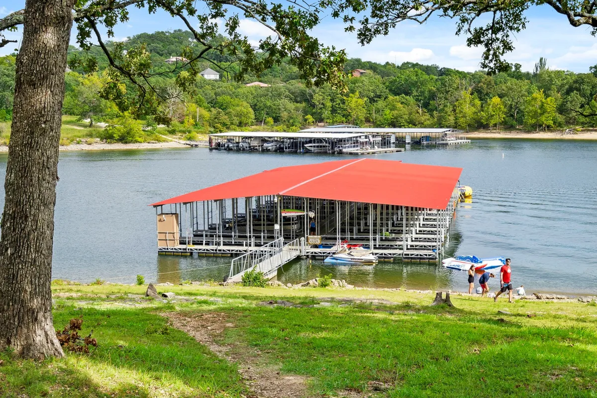 A red-roofed boat dock extends over the water, with people strolling by the lakeshore amid lush greenery. The scene feels open and inviting, with grassy areas ideal for walking and playing, and the lake offering scenic views.