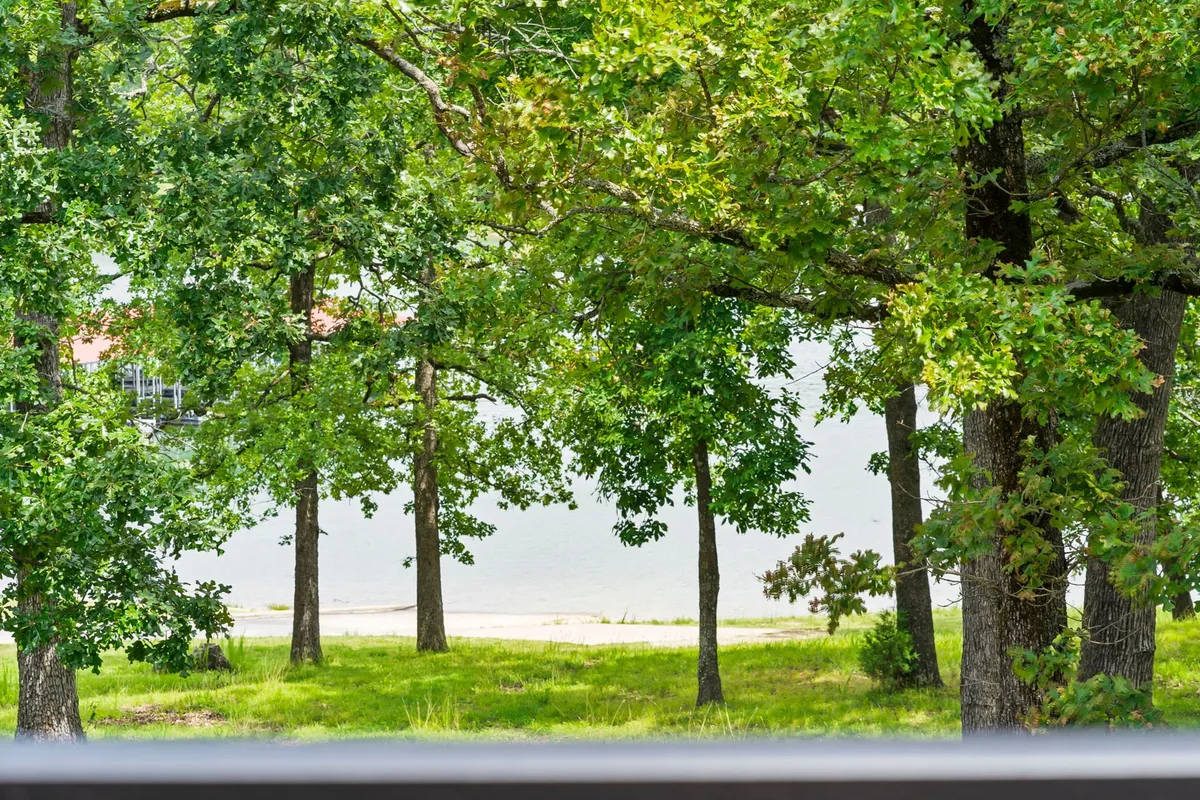 A serene view of lush green trees bordering calm water with a sandy shore in the foreground, ideal for picnicking, fishing, or boating. The tranquil lake setting captures the essence of lakeside leisure and outdoor group activities.
