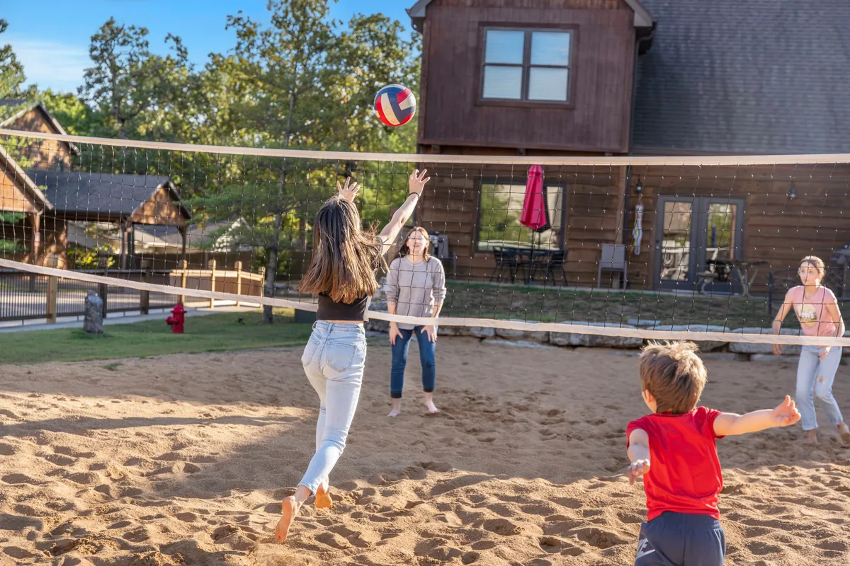 This shot captures families and children actively playing volleyball on a sand court, with both adults and children participating. The vacation home is visible in the background, situating the activity within the resort context.