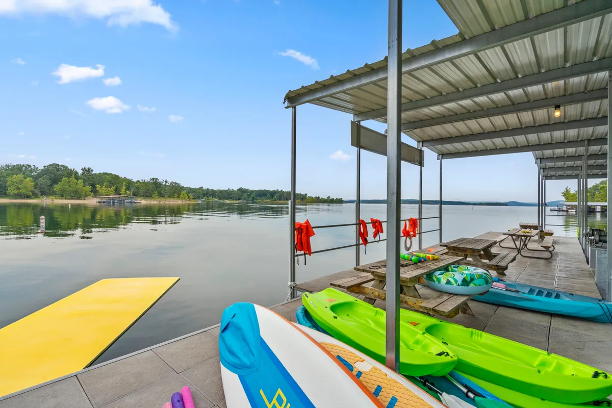 Colorful kayaks rest on a yellow floating dock beside calm water, with a picnic table and life jackets nearby. The area is inviting for paddling, sunbathing, swimming, and relaxing, all in a family-friendly lakeside setting.