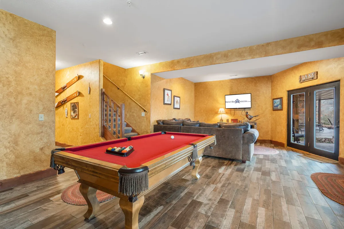 A spacious, inviting game room featuring a pool table, comfortable sofa, television, and a modern staircase. The room radiates a casual, social mood, perfect for friendly competition, movie nights, and group relaxation.