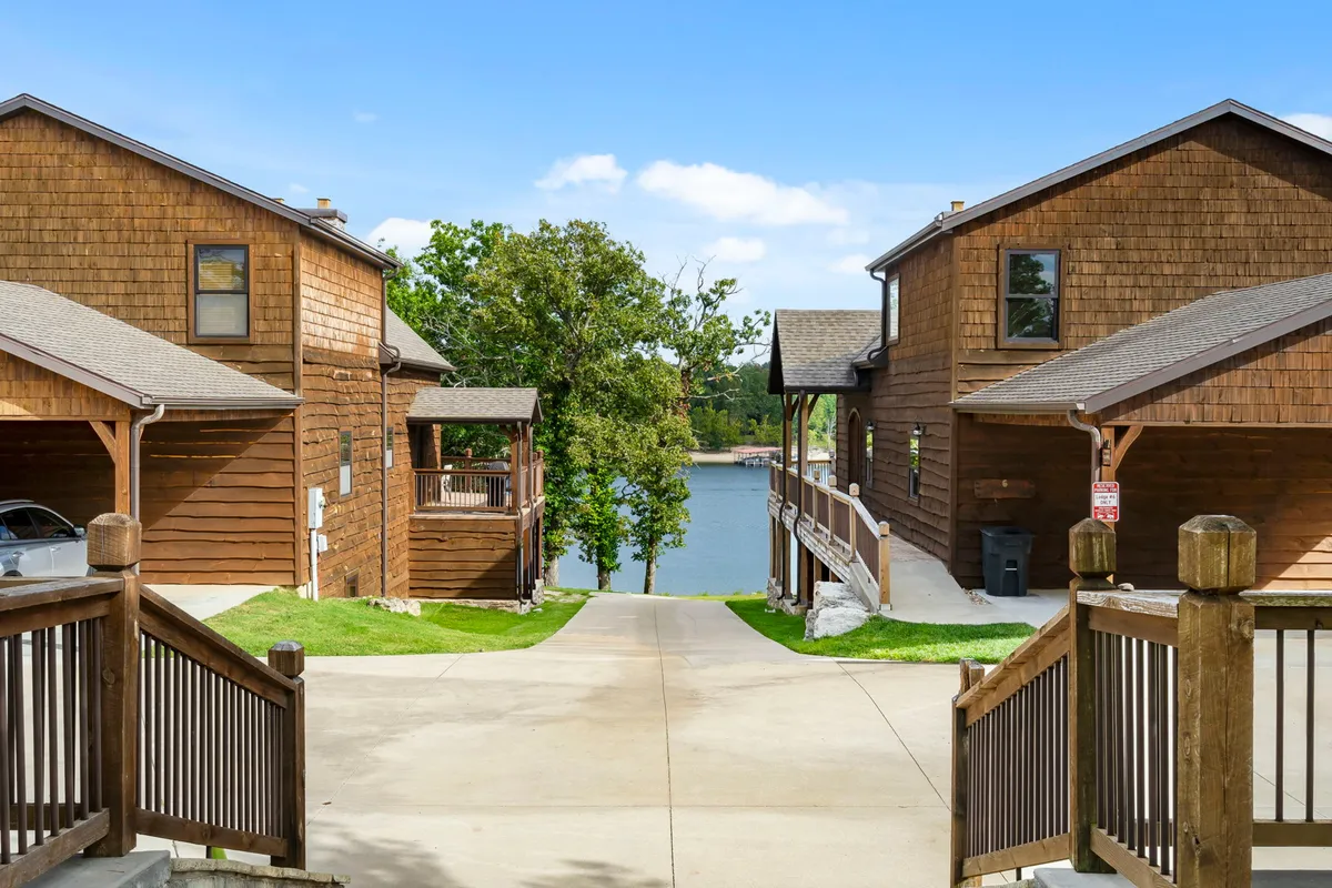 Wooden cabins line a tranquil lakefront with a pathway and abundant greenery. The wide, open space connects the cabins to the water, creating an inviting area for walking, relaxing, and enjoying nature.