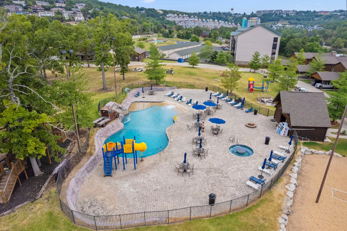 This image showcases a vibrant outdoor area featuring a swimming pool, hot tub, playground equipment, and lounge chairs under umbrellas. The pool area is inviting and ideal for family gatherings, relaxation, and play. The playground is visible, emphasizing amenities for children.