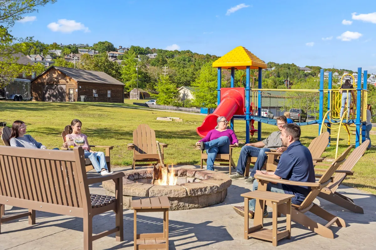 A circular fire pit is surrounded by comfortable seating and playground equipment, set within a green lawn. The scene captures children playing on the playground and families gathering around the fire pit, creating a communal and welcoming atmosphere.