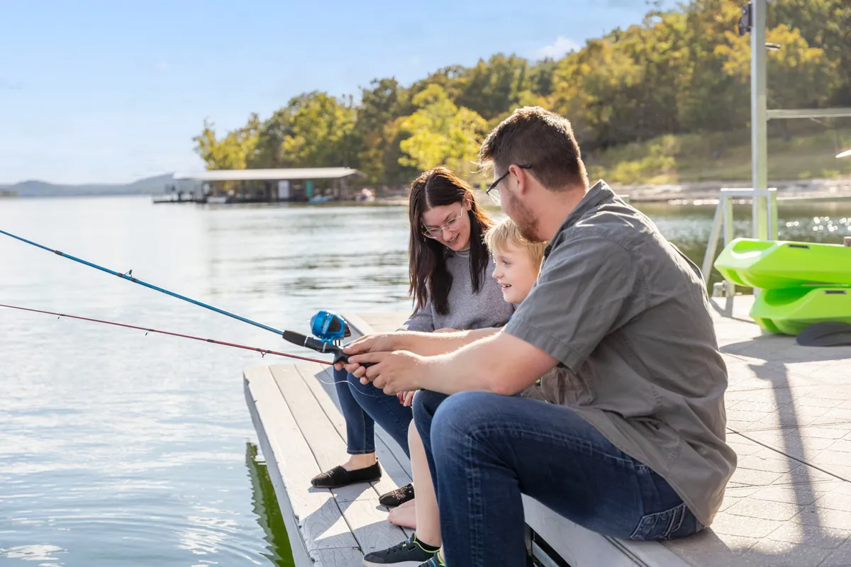 A family is gathered on a dock by the calm lake, with fishing rods in hand and green trees surrounding the water. The scene exudes togetherness, with children and adults enjoying fishing, relaxing by the water, and sharing a picnic.