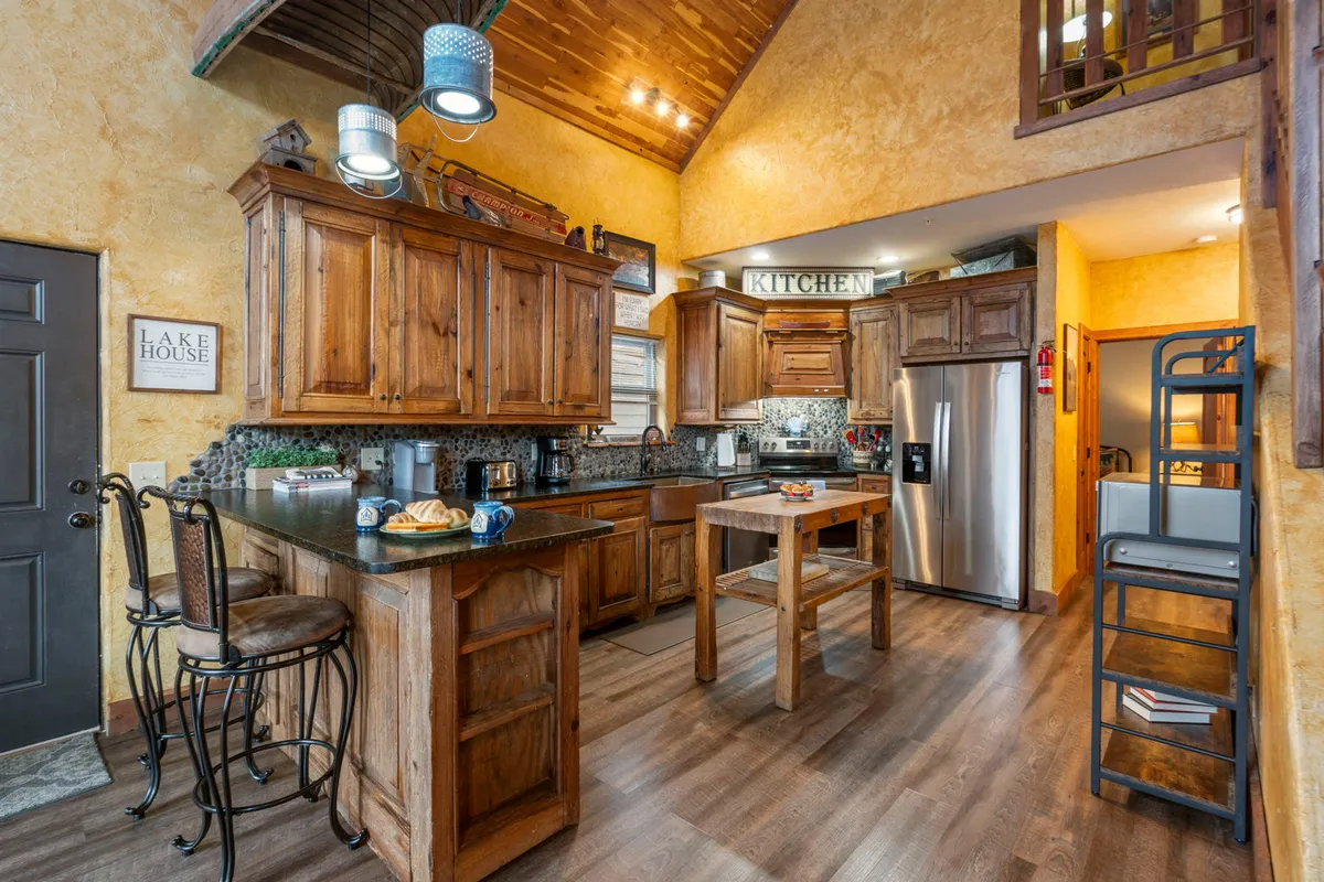 The image showcases a warm and inviting kitchen with elegant wooden cabinets, a spacious kitchen island surrounded by bar stools, and tasteful decorative signage. The setting is perfect for cooking, gathering, and socializing, creating an atmosphere ideal for family meals, entertaining friends, or enjoying casual brunches together.